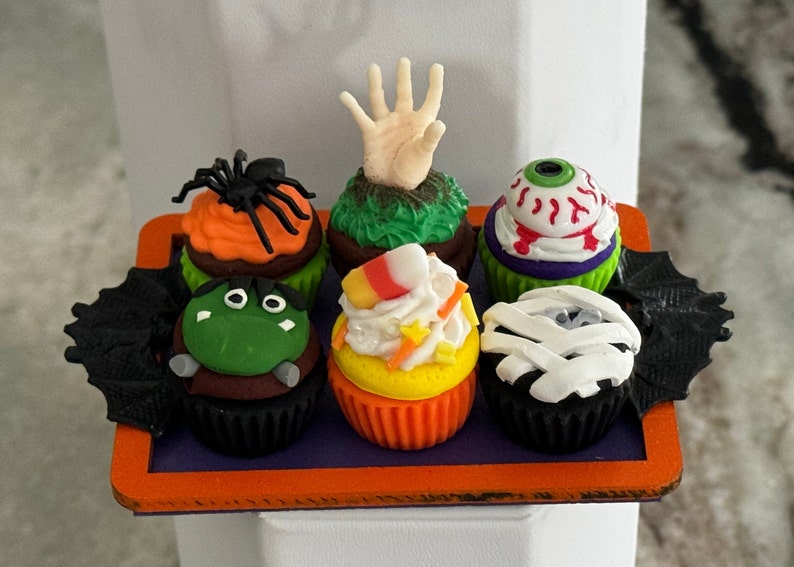Op de afbeelding: Zes Halloween-cupcakes versierd met verschillende griezelige figuren en ontwerpen. De cupcakes zijn gerangschikt in een ronde oranje schaal met zwarte vleermuisvleugels aan beide kanten.