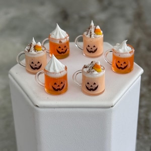 May include: Six miniature clear glass mugs filled with orange liquid, each featuring a black jack-o'-lantern face. The mugs are topped with white whipped cream, dark brown sprinkles, and a single piece of yellow, orange, and white candy corn.