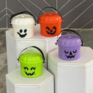May include: Four small Halloween buckets in white, orange, purple, and green, each with a black handle. The buckets feature jack-o'-lantern faces and a ghost face. The buckets are displayed on white pedestals.