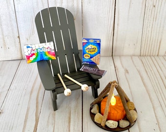 May include: A miniature camping scene with a wooden Adirondack chair, a small fire pit with a lit candle, and camping supplies like marshmallows, graham crackers, and chocolate. The supplies are labeled "Jet-Puffed", "Honey Maid", and "Hershey's".