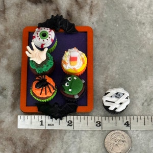 Op de afbeelding: Een set van zes Halloween-thema cupcakes op een paarse en oranje dienblad. De cupcakes zijn versierd met een oog, een hand, een ma&iuml;s, een spin, een monster en een mummie.