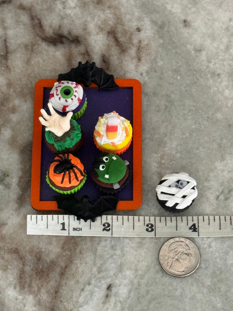 Op de afbeelding: Een set van zes Halloween-thema cupcakes op een paarse en oranje dienblad. De cupcakes zijn versierd met een oog, een hand, een ma&iuml;s, een spin, een monster en een mummie.