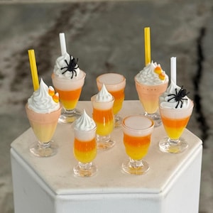 May include: A collection of miniature glass dessert cups filled with layered yellow, orange, and white candy corn-colored liquid. Some cups are topped with white whipped cream, black plastic spiders, yellow straws, or candy corn pieces.