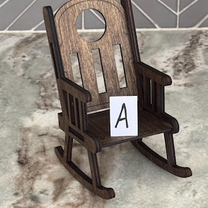 Peut inclure: Une chaise ber&ccedil;ante miniature en bois avec une finition marron fonc&eacute;. La chaise a un dessus arrondi avec une d&eacute;coupe circulaire et des lattes verticales. Une petite carte blanche avec la lettre "A" est plac&eacute;e sur le si&egrave;ge.