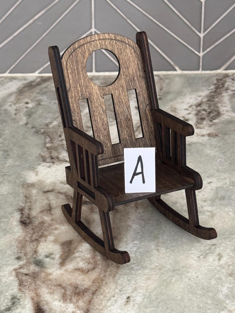 Peut inclure: Une chaise ber&ccedil;ante miniature en bois avec une finition marron fonc&eacute;. La chaise a un dessus arrondi avec une d&eacute;coupe circulaire et des lattes verticales. Une petite carte blanche avec la lettre "A" est plac&eacute;e sur le si&egrave;ge.
