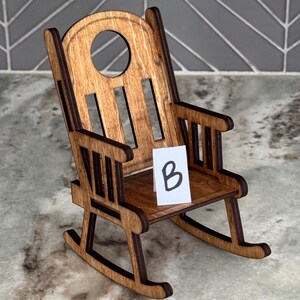 Peut inclure: Une chaise ber&ccedil;ante miniature en bois avec une finition marron fonc&eacute;. La chaise a un dossier arrondi avec une d&eacute;coupe circulaire et une petite assise. Une carte blanche avec la lettre "B" est sur le si&egrave;ge.
