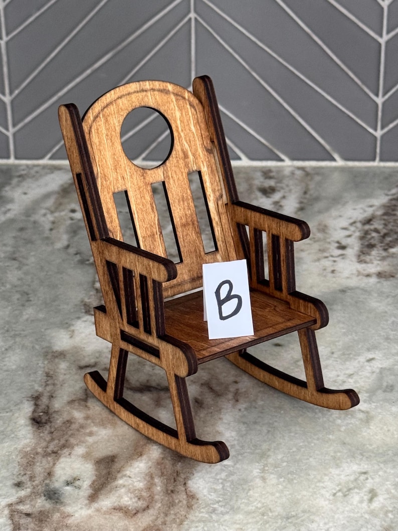 Peut inclure: Une chaise ber&ccedil;ante miniature en bois avec une finition marron fonc&eacute;. La chaise a un dossier arrondi avec une d&eacute;coupe circulaire et une petite assise. Une carte blanche avec la lettre "B" est sur le si&egrave;ge.