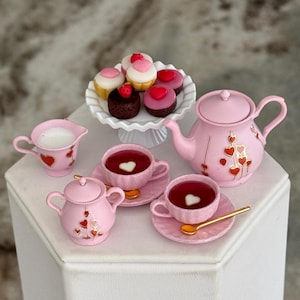 May include: A miniature pink tea set with heart accents, including a teapot, creamer, sugar bowl, and two teacups with saucers. A white tiered plate holds miniature cupcakes with heart and strawberry toppings. A golden spoon rests on a saucer.