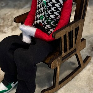 Peut inclure: Une poup&eacute;e v&ecirc;tue d'une chemise rouge, d'un pantalon noir et d'un gilet pied-de-poule est assise dans un rocking-chair en bois. Les chaussures de la poup&eacute;e sont blanches et vertes. La chaise est en bois brun fonc&eacute;.
