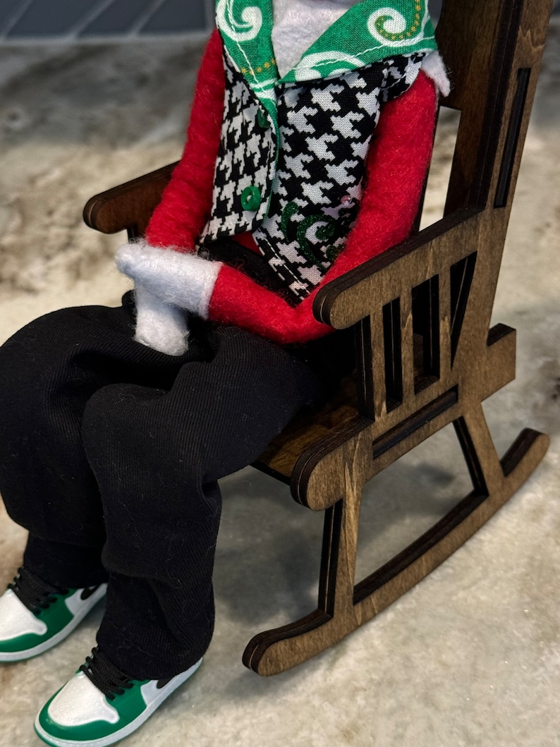 Peut inclure: Une poup&eacute;e v&ecirc;tue d'une chemise rouge, d'un pantalon noir et d'un gilet pied-de-poule est assise dans un rocking-chair en bois. Les chaussures de la poup&eacute;e sont blanches et vertes. La chaise est en bois brun fonc&eacute;.
