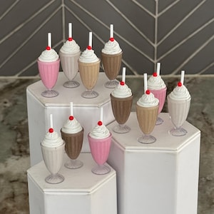 May include: A set of ten miniature milkshakes with whipped cream and a red cherry on top. The milkshakes are in clear plastic cups with straws and are arranged on two white hexagonal tiered stands.