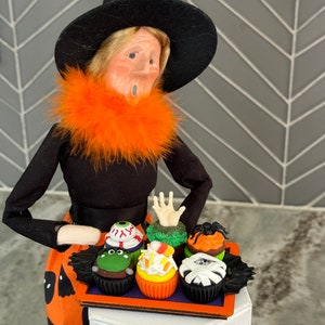 Op de afbeelding: Een heksenpop met een zwarte hoed en een oranje bontkraag houdt een dienblad met Halloween-cupcakes vast. De cupcakes zijn versierd met groene, oranje, witte en zwarte frosting en hebben verschillende Halloween-versieringen, waaronder een oog, een hand, een spin en een mummie.