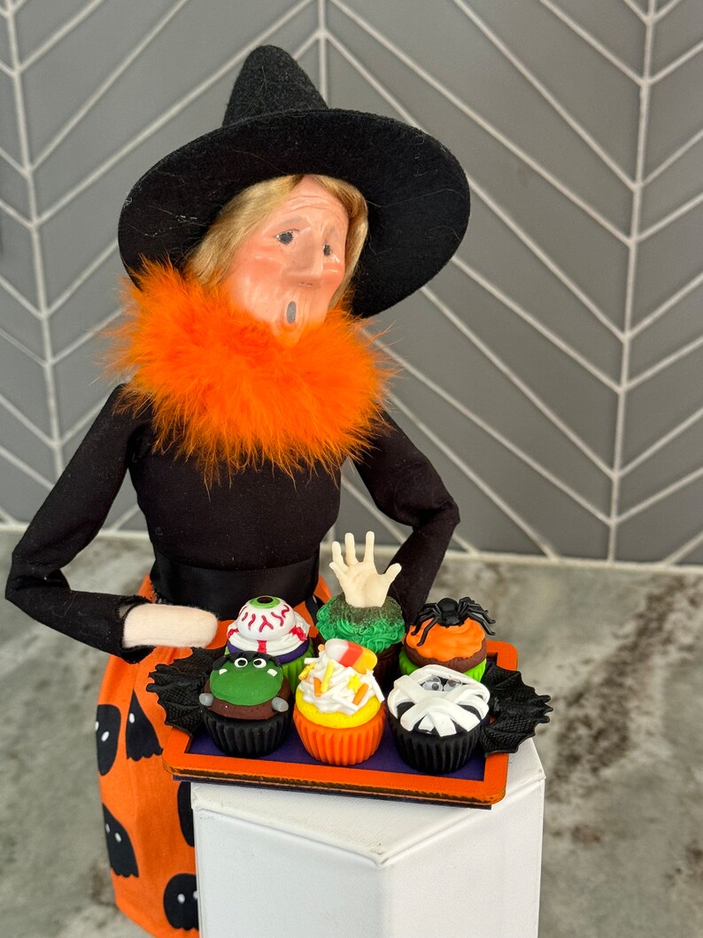 Op de afbeelding: Een heksenpop met een zwarte hoed en een oranje bontkraag houdt een dienblad met Halloween-cupcakes vast. De cupcakes zijn versierd met groene, oranje, witte en zwarte frosting en hebben verschillende Halloween-versieringen, waaronder een oog, een hand, een spin en een mummie.