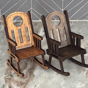 Peut inclure: Deux petites chaises &agrave; bascule en bois. L'une est marron clair, l'autre est marron fonc&eacute;. Chaque chaise a un dossier arrondi avec une d&eacute;coupe circulaire et des lattes verticales. Les chaises sont sur une surface claire.