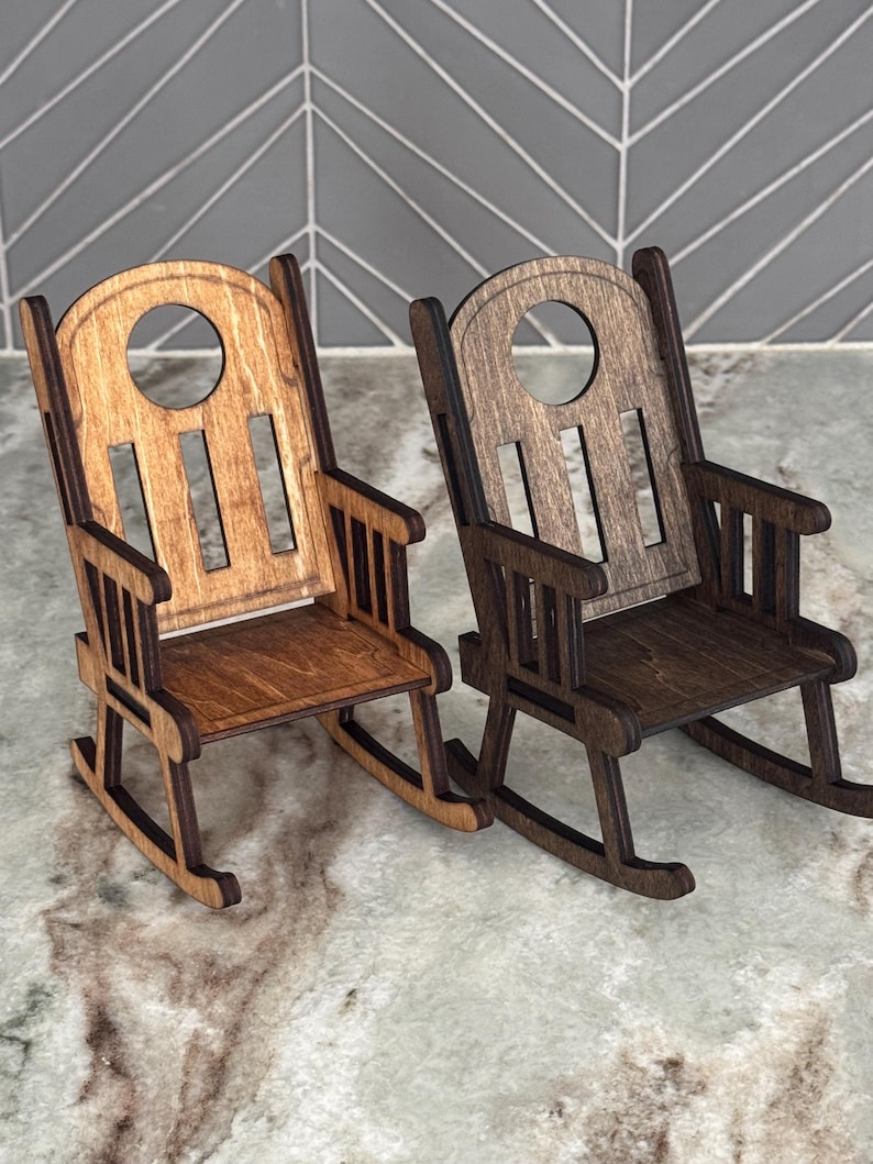 Peut inclure: Deux petites chaises &agrave; bascule en bois. L'une est marron clair, l'autre est marron fonc&eacute;. Chaque chaise a un dossier arrondi avec une d&eacute;coupe circulaire et des lattes verticales. Les chaises sont sur une surface claire.