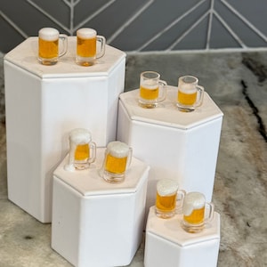 May include: A collection of eight miniature clear plastic beer mugs filled with a golden liquid and topped with white foam. The mugs are arranged in pairs on four white hexagonal pedestals of varying heights, creating a tiered display.