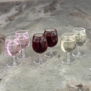 May include: A set of six miniature clear glass goblets arranged in pairs, featuring two filled with light pink liquid, two with deep red liquid, and two with clear liquid. Each small stemware piece has a rounded bowl and a circular base.
