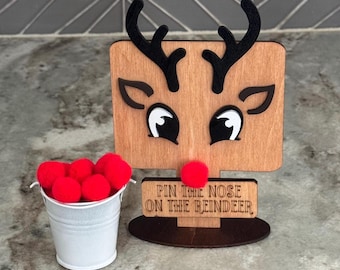 May include: A wooden reindeer game board featuring black antlers, black and white eyes, and a red pom-pom nose, with the text "PIN THE NOSE ON THE REINDEER" engraved on a rectangular plaque, accompanied by a small white bucket filled with red pom-poms.