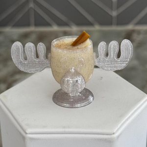 May include: A clear glass mug shaped like a moose head with antlers, filled with a white eggnog-like drink and a cinnamon stick garnish.