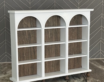 May include: White wooden bookcase with three arched sections and four shelves in each section. The shelves have a wood-grain pattern.