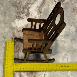 Peut inclure: Une chaise ber&ccedil;ante miniature en bois avec une finition marron fonc&eacute;. La chaise a un dossier &agrave; lattes avec une d&eacute;coupe ovale et mesure environ 10 cm de long. La chaise est plac&eacute;e sur une surface de couleur claire.