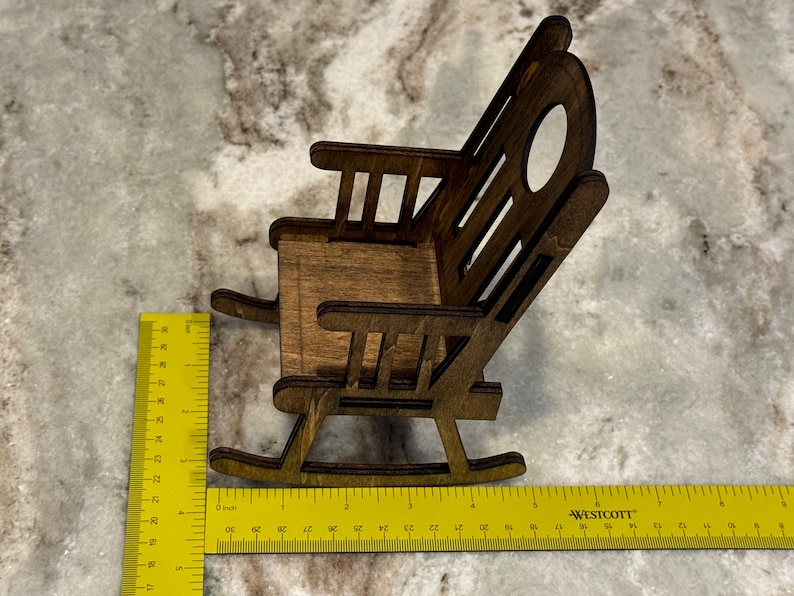 Peut inclure: Une chaise ber&ccedil;ante miniature en bois avec une finition marron fonc&eacute;. La chaise a un dossier &agrave; lattes avec une d&eacute;coupe ovale et mesure environ 10 cm de long. La chaise est plac&eacute;e sur une surface de couleur claire.