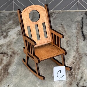 Peut inclure: Une petite chaise &agrave; bascule en bois de couleur marron clair. Le dossier de la chaise est arrondi avec une d&eacute;coupe circulaire et des lattes verticales. L'assise et les accoudoirs sont &eacute;galement en bois. Une petite carte blanche avec la lettre "C" est devant la chaise.