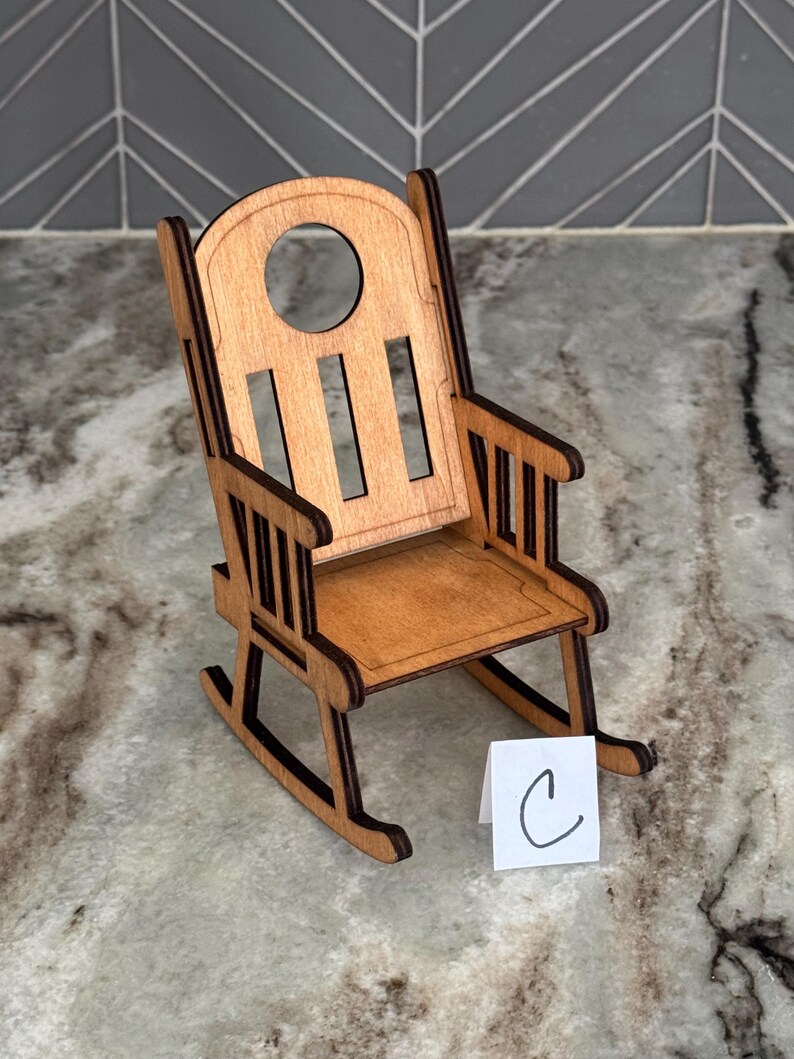 Peut inclure: Une petite chaise &agrave; bascule en bois de couleur marron clair. Le dossier de la chaise est arrondi avec une d&eacute;coupe circulaire et des lattes verticales. L'assise et les accoudoirs sont &eacute;galement en bois. Une petite carte blanche avec la lettre "C" est devant la chaise.