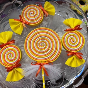 May include: A selection of spiral-patterned lollipops in yellow, orange, and white. Each sweet is decorated with a yellow bow and a red ribbon. One lollipop features a white tulle skirt and a yellow stick, presented on a glass plate.