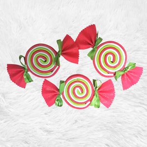 May include: Four handmade candy-shaped ornaments with green and red stripes. Each ornament is attached to a pink bow with green ribbon.