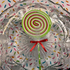 May include: A green, white, and red swirl lollipop with a white tulle bow and red ribbon on a green and white striped stick. The lollipop is sitting on a clear glass plate with a colorful confetti pattern.