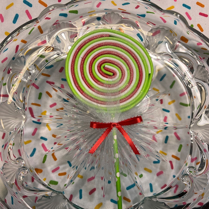 May include: A green, white, and red swirl lollipop with a white tulle bow and red ribbon on a green and white striped stick. The lollipop is sitting on a clear glass plate with a colorful confetti pattern.