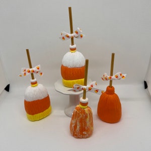 May include: Four Halloween decorative brooms in the shape of candy corn. The brooms feature white, orange, and yellow sections. Each has a wooden handle and a candy corn ribbon bow. One broom is displayed on a white pedestal.