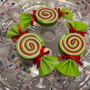 May include: Four green and white candy cane ornaments with red bows. The ornaments are on a clear glass plate with a colorful confetti pattern.