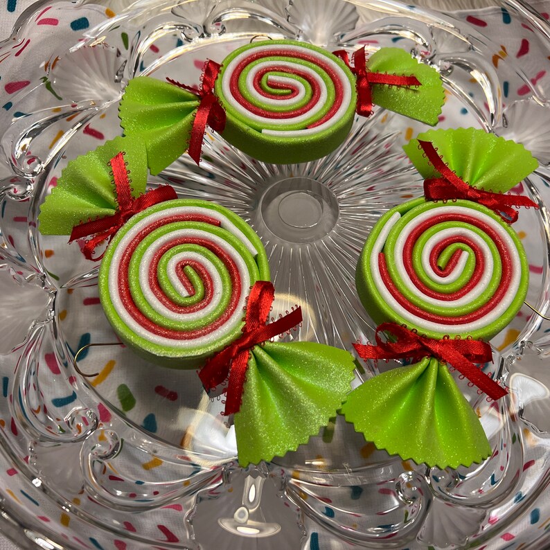 May include: Four green and white candy cane ornaments with red bows. The ornaments are on a clear glass plate with a colorful confetti pattern.