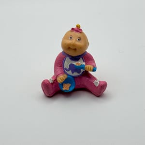 May include: A Cabbage Patch Kids figurine of a baby in a pink outfit, bib, and a blue bowl and spoon. The baby has a pink bow on its head and is sitting down. The bib has a purple elephant design.