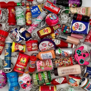 May include: A collection of miniature keychains featuring various food and beverage brands. The keychains include items like ketchup bottles, soda cans, and candy, all attached to silver keyrings. The items are in various colors, including red, blue, and yellow.