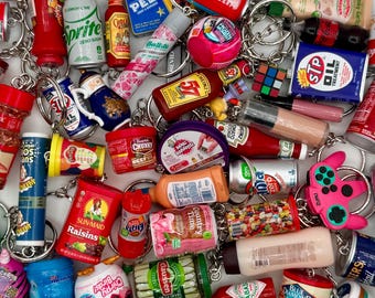 May include: A collection of miniature keychains featuring various food and beverage brands. The keychains include items like ketchup bottles, soda cans, and candy, all attached to silver keyrings. The items are in various colors, including red, blue, and yellow.