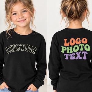 May include: A black long-sleeve shirt featuring the word CUSTOM in a yellow arched font on the front. The back displays the words LOGO PHOTO TEXT in a stacked, wavy, multi-coloured font with orange, green, and purple lettering on a black background.