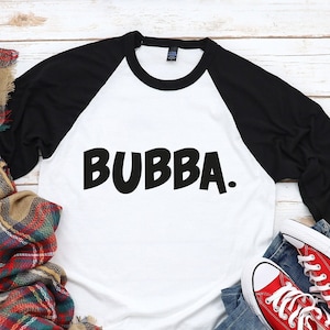 May include: A white and black baseball tee shirt with the word "BUBBA." printed in black on the front. The shirt is laying on a white wood surface with a red and black plaid scarf, a red mug, and red and white sneakers.