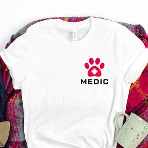 May include: White t-shirt with a red paw print with a white plus sign in the center and the word "MEDIC" in black letters below the paw print.
