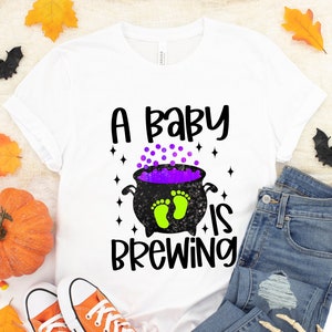 May include: A white t-shirt with a black and green graphic of a cauldron with a purple liquid and two green footprints inside. The text "A baby is brewing" is written in black above the cauldron.