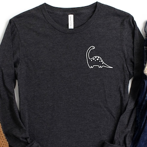 Peut inclure: Un t-shirt à manches longues gris foncé avec un motif de dinosaure blanc sur le devant. Le t-shirt est associé à un jean bleu, un bonnet en tricot beige et des ballerines tissées brunes.