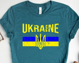 Pode incluir: Uma camiseta azul turquesa com a palavra "UKRAINE" em letras amarelas e uma bandeira ucraniana amarela e azul com um tridente.