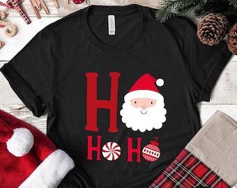 May include: Black t-shirt with a red "Ho Ho Ho" graphic featuring a cartoon Santa Claus, a candy cane, and a bauble. The shirt is surrounded by Christmas decorations, including a Santa hat, a stocking, and a Christmas garland.