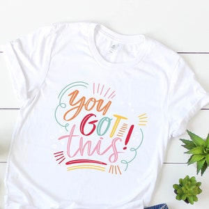 Op de afbeelding: Wit T-shirt met de tekst "you got this!" in kleurrijke letters op een witte houten achtergrond. Het shirt wordt gecombineerd met een spijkerbroekje en sandalen.