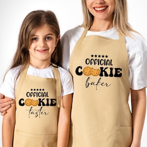May include: Two people wearing tan aprons with the text "Official Cookie Baker" and "Official Cookie Taster" on them. The aprons have pockets and are made of a soft, comfortable fabric.