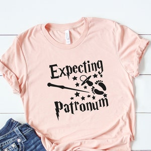 May include: A pink t-shirt with a black graphic that says "Expecting Patronum" with a wand, a pacifier, and baby footprints.