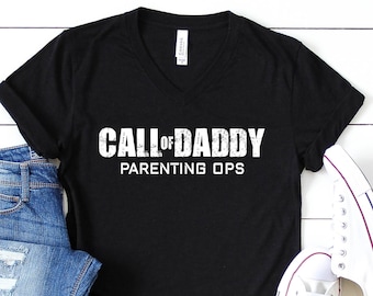 May include: Black V-neck t-shirt with the text "CALL OF DADDY PARENTING OPS" in white. The shirt is paired with blue denim jeans and white sneakers. The image is taken on a white wooden surface.