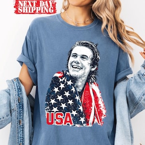 May include: Blue t-shirt featuring a graphic of a smiling person draped in an American flag. The word "USA" is printed in red below the image. The shirt has rolled-up sleeves.